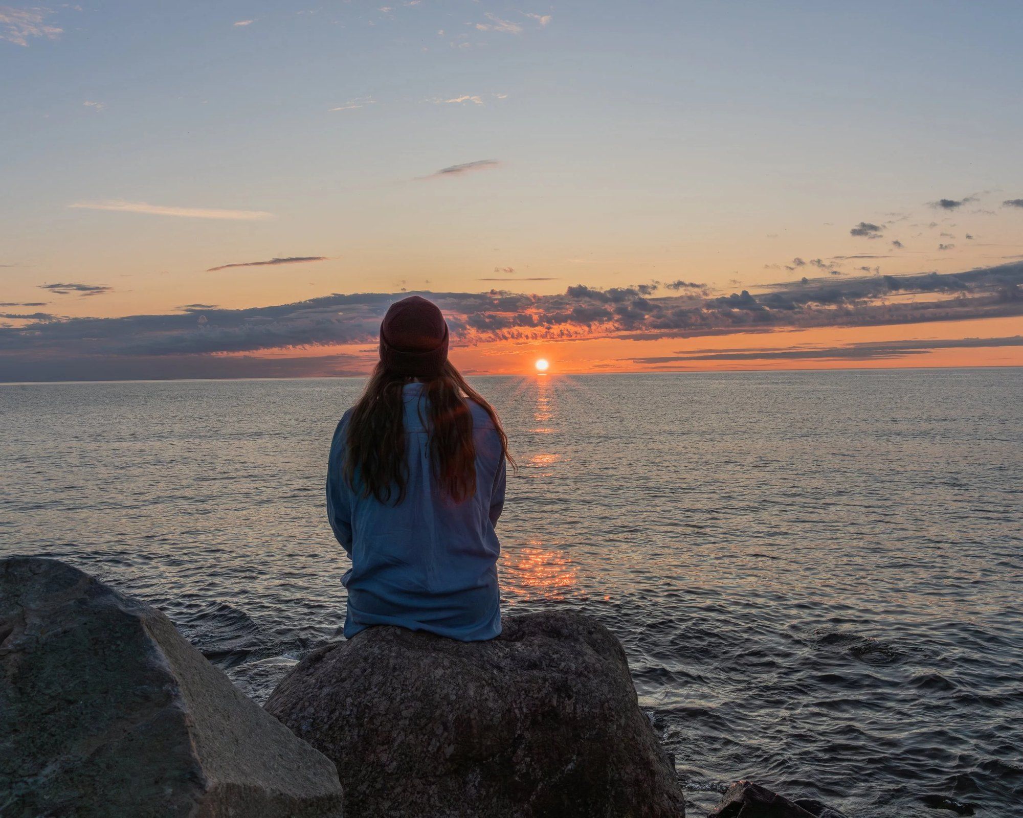 Nainen istuu rantakivellä ja katsoo auringonlaskua meren yllä