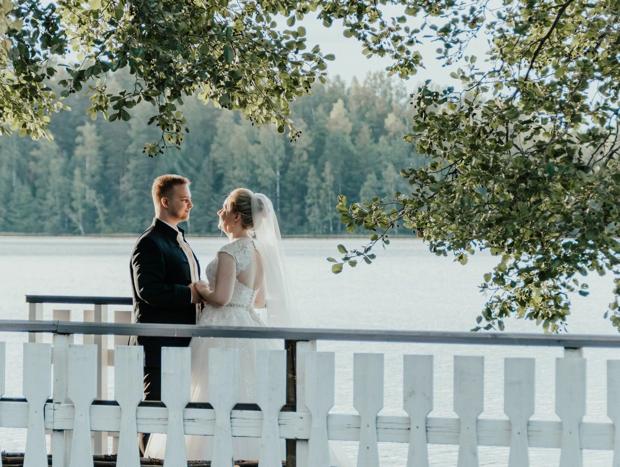 Hääpari seisoo valkoisen kaiteen äärellä järven rannalla